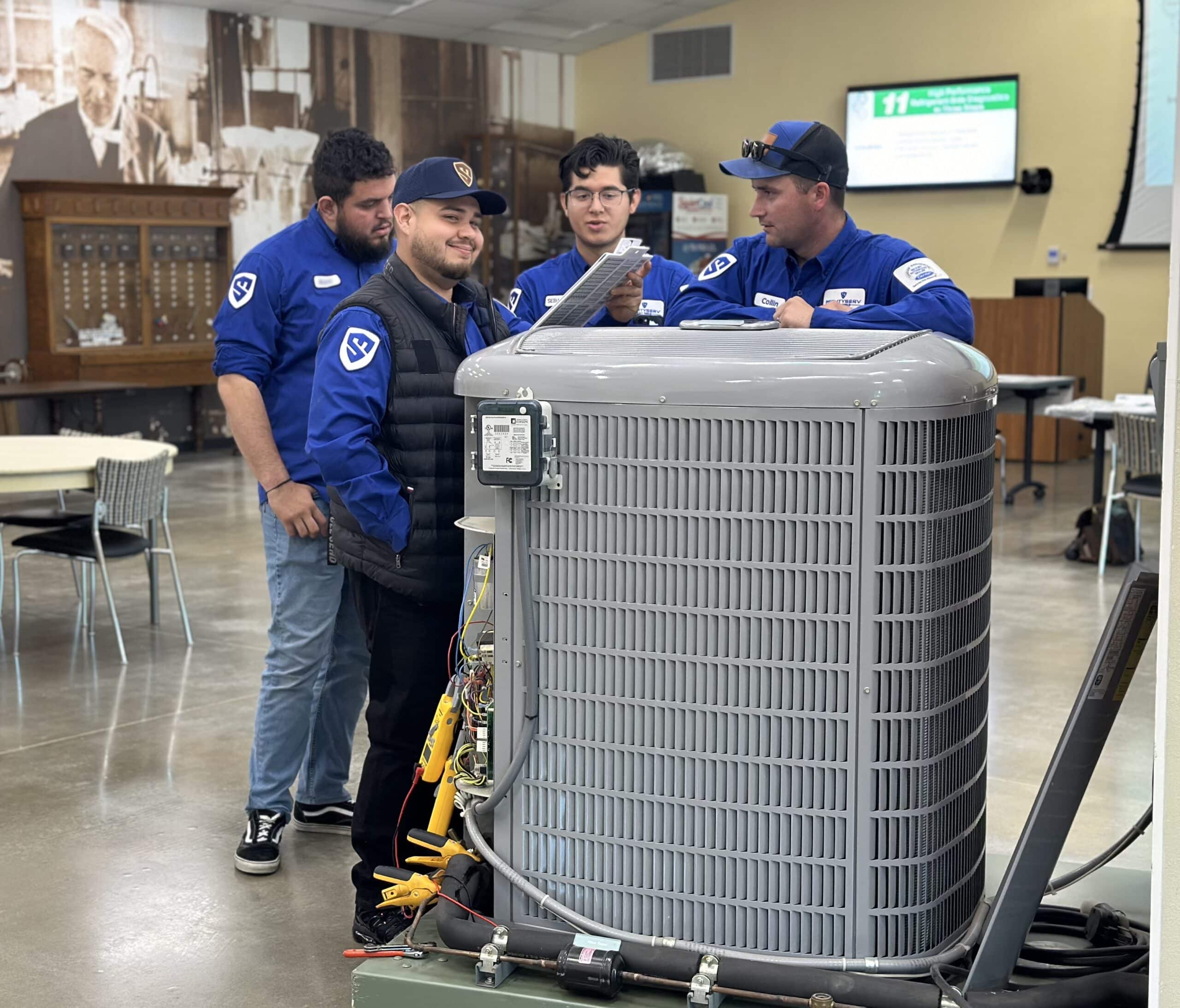 Four MightyServ HVAC technicians in blue uniforms gathered around an open air conditioning condenser unit, reviewing a checklist during a technical training exercise