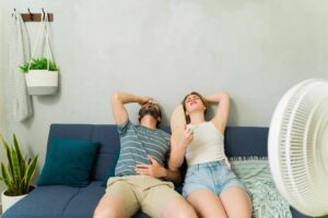 A couple leaning back on a couch with hands raised, sitting in front of a large fan to stay cool.