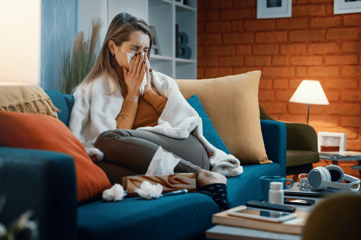 A woman on a blue couch blowing her nose into a tissue, looking congested and uncomfortable.